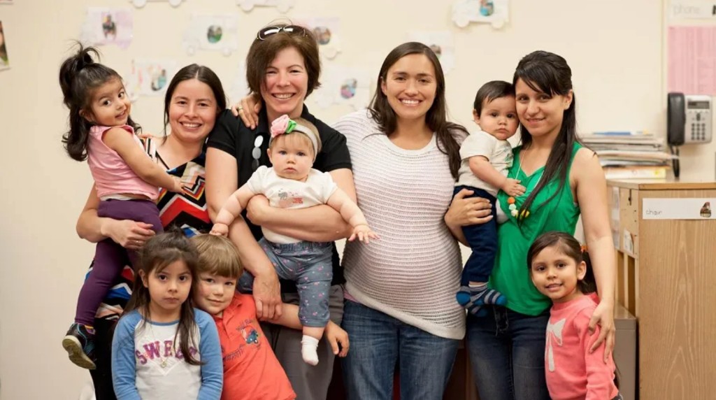 Julie sonriendo con un grupo de madres y niños pequeños en un ambiente comunitario.